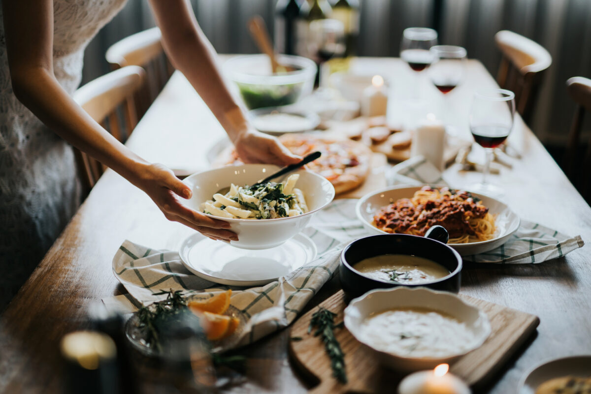 table being set for dinner, woman holding bowl of pasta above table