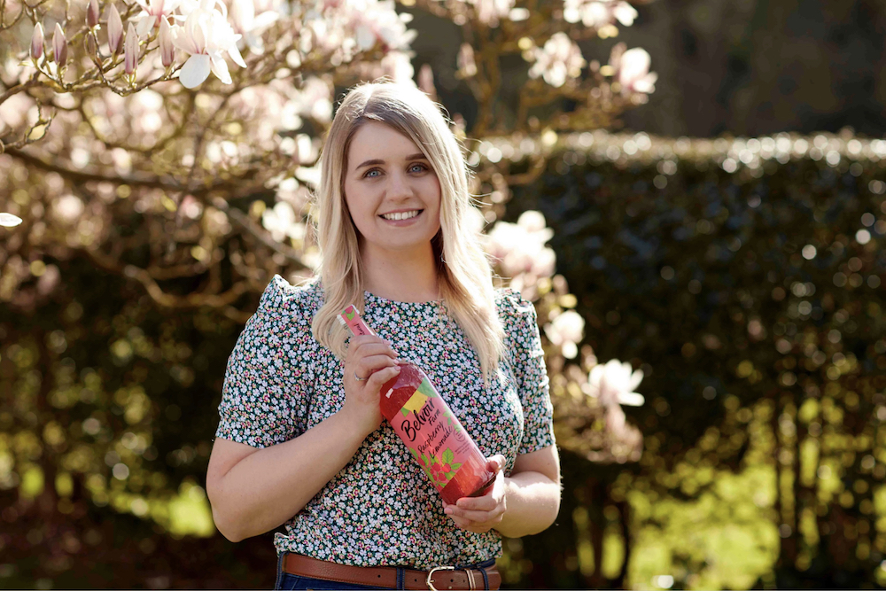 Jessica Pinnick, Brand Manager at Belvoir Farm, holding a red bottle smiling in the countryside