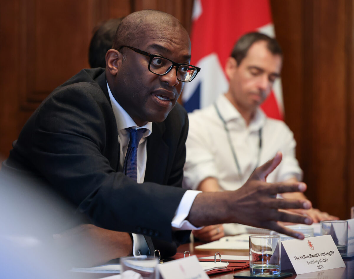 kwasi kwarteng reaching across a table with a man and union jack in the back ground