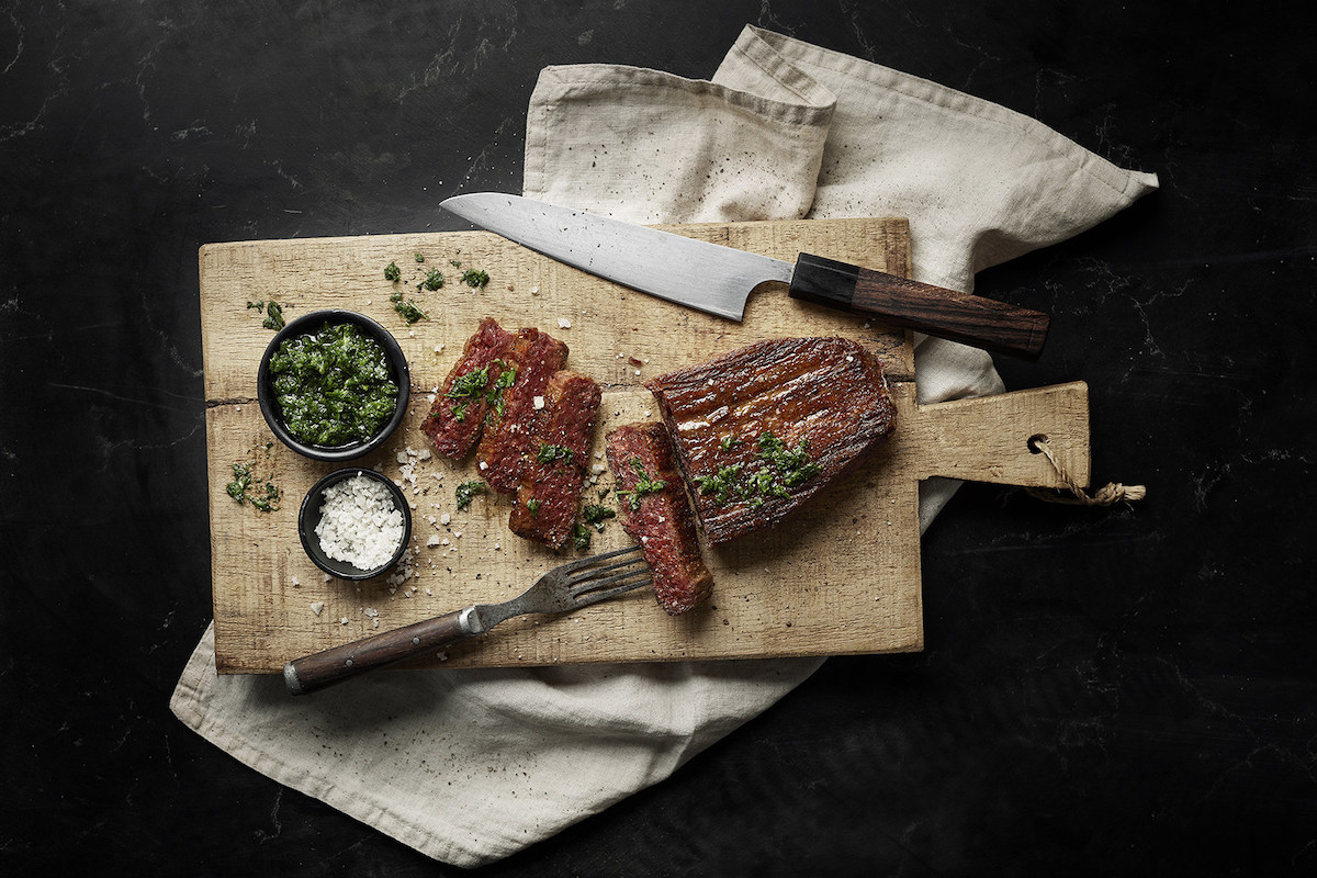 chopped up 3D-printed plant-based meat on wooden board with large knife and fork, bowl of parsely and salt on black background with napkin