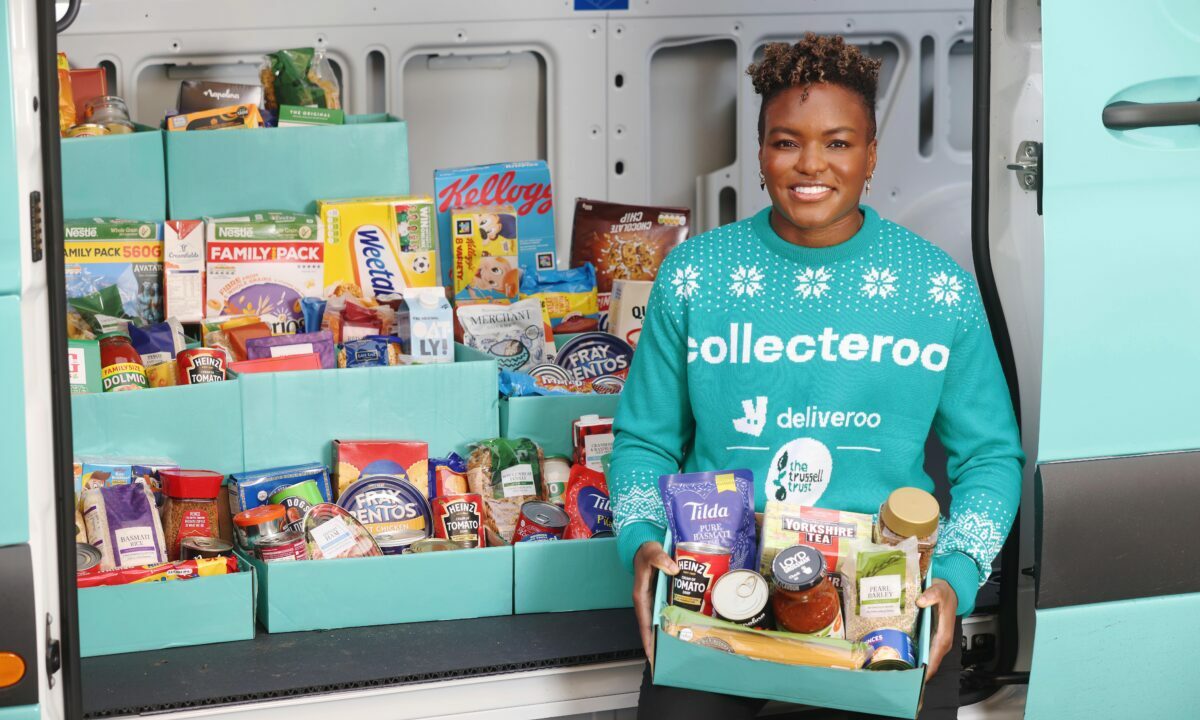 Black woman wearing blue jumper holding box of dry and tinned food items in front of other items laid out in blue boxes.