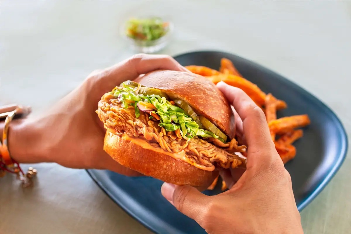 Fried chicken sandwich in bread bun held in hands above blue plate with chips