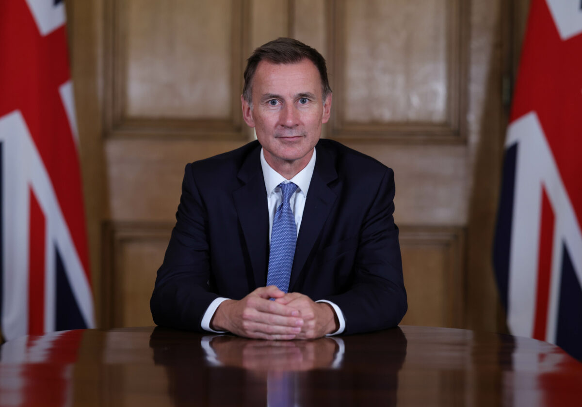 UK Chancellor of the Exchequer Jeremy Hunt, man sitting at table looking face forward inbetween two Union Jack flags, wooden wall background