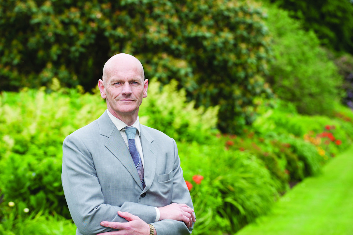 Image of bald man in grey suit in front of greenery background.