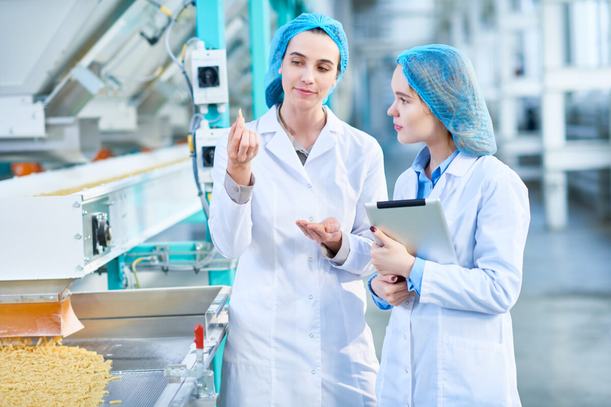 Two young female factory workers in factory plant looking at piece of pasta