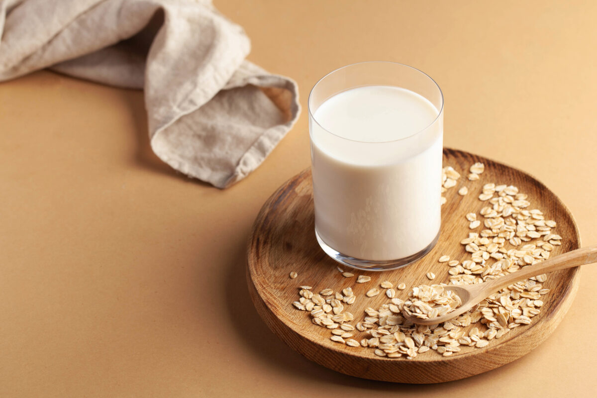 glass of oat milk on wooden plate with oats and wooden spoon, beige background with cloth