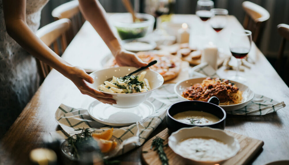 table being set for dinner, woman holding bowl of pasta above table