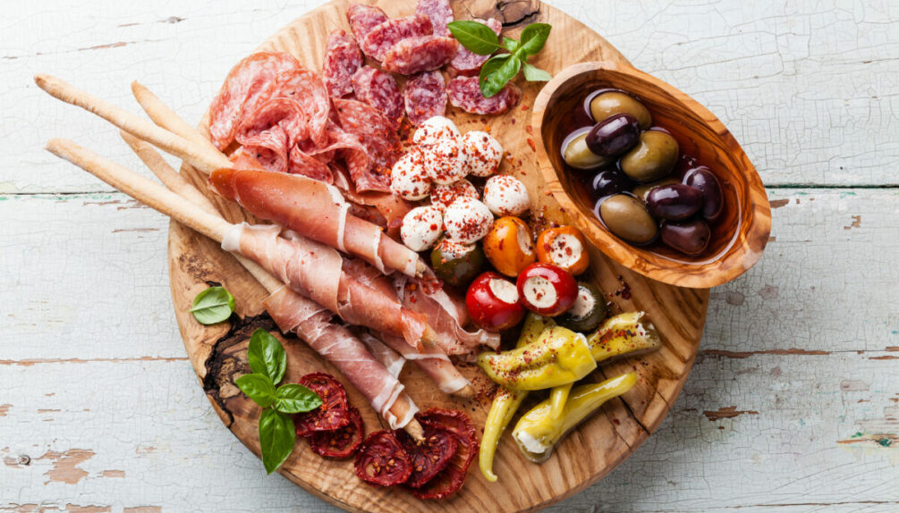 board of various italian meats and cheeses, as well as olives and breadsticks, against a white background