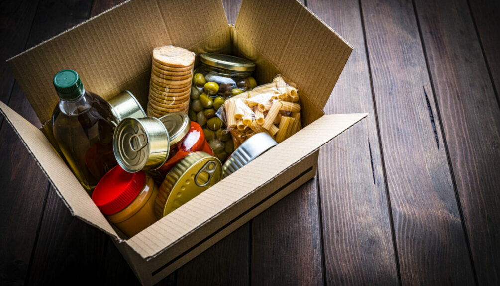 High angle view of a cardboard box filled with multicolored non-perishable canned goods, conserves, sauces and oils shot on dark wooden background