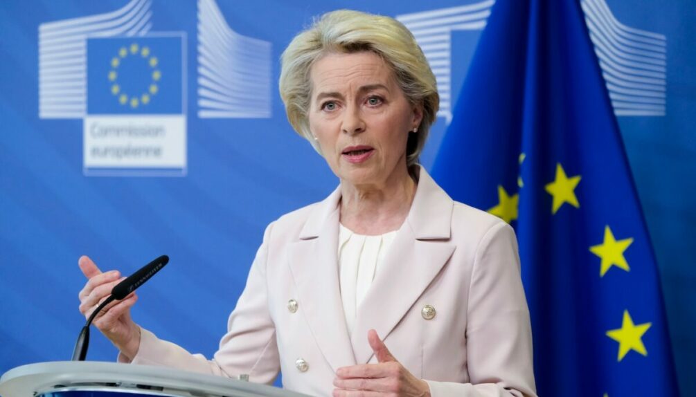 EU Commission President Ursula von der Leyen gives speech on podium in front of blue background and blue and yellow EU flag