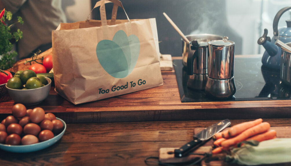 too good to go bag food waste innovation on the kitchen countertop, next to a coffee pot and surrounded by various produce including carrots and apples