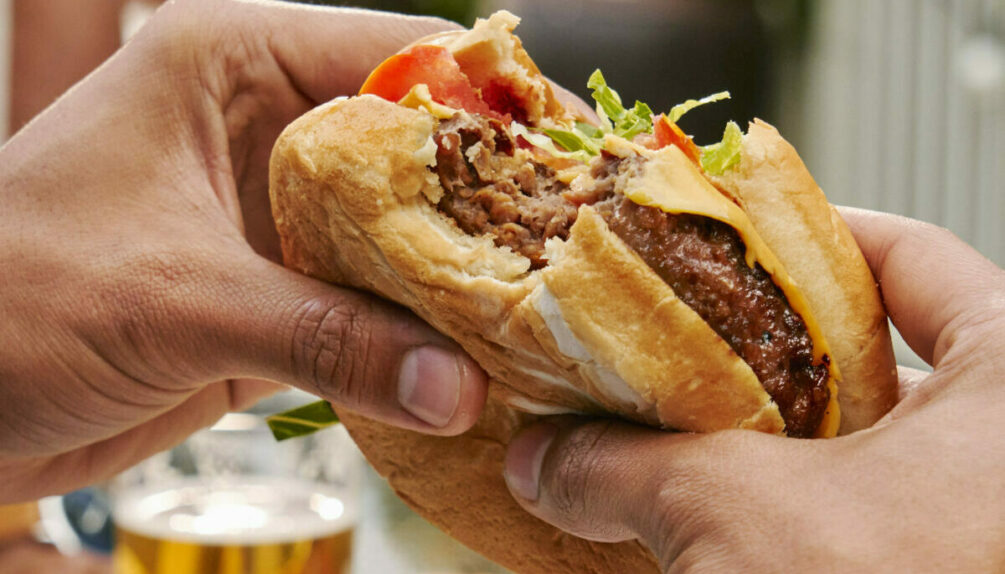 plant-based burger patty in bun with cheese, lettuce and tomato in person's hands, in front of beer in glass in background