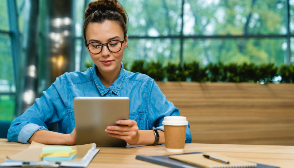young woman with glasses and blue shirt sits at wooden table looking at tablet in glass window room with plastic coffee cup and notepads