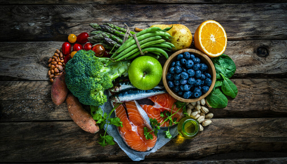 pile of food on wooden table including apples, broccoli, green beans, salmon and oranges
