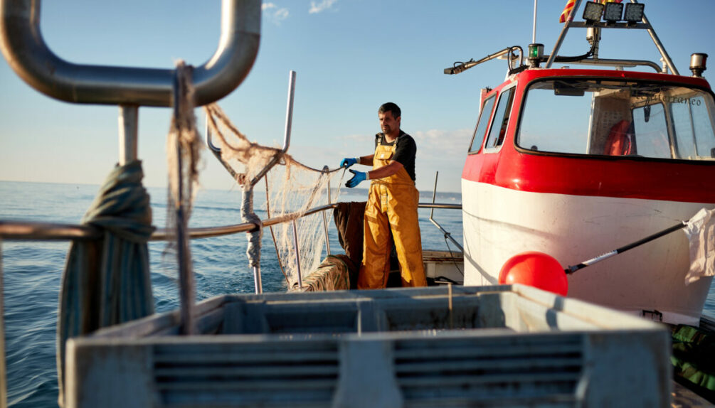 Front view of Caucasian man in early 30s wearing protective workwear and standing at stern working with trawl nets at sea in early morning