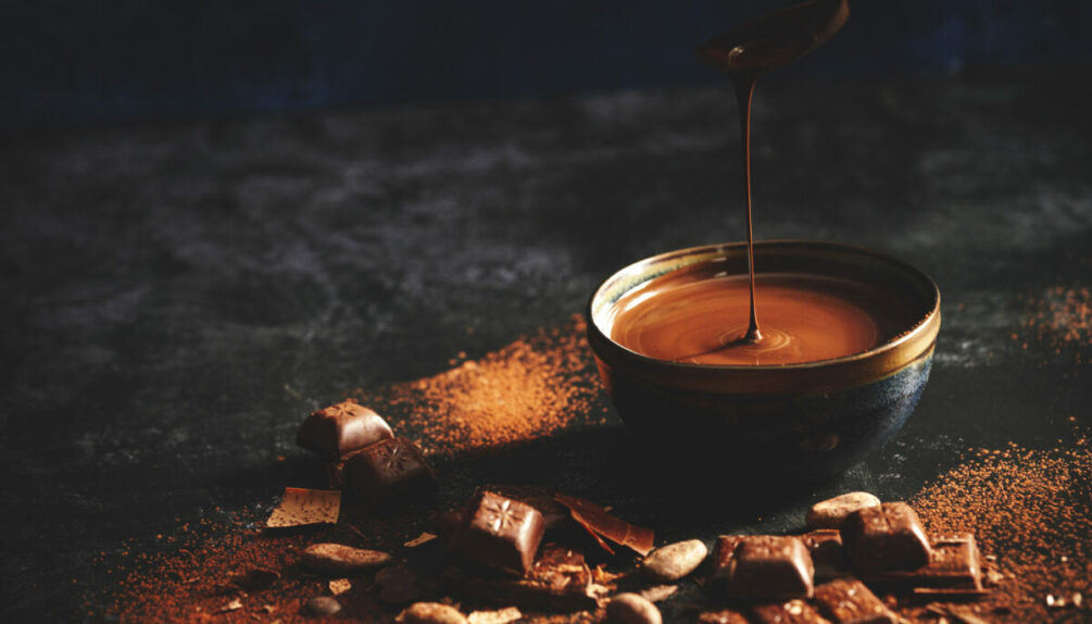 melting chocolate in bowl surrounded by chocolate squares, cocoa powder, and nuts on dark background