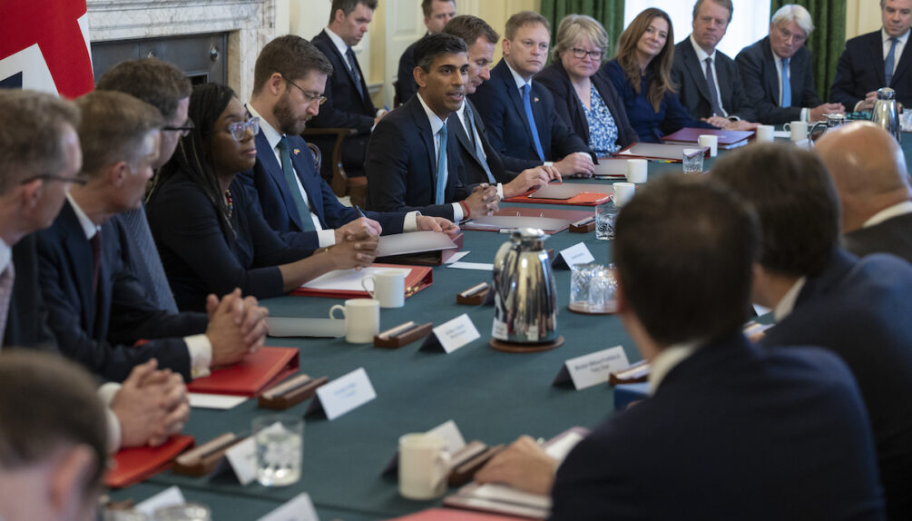 UK Prime Minister talking with colleagues in cabinet meeting, all sat on long green table