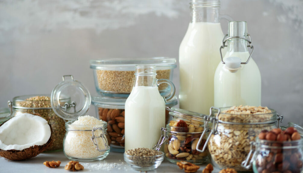 Glass bottles of vegan plant milk and almonds, nuts, coconut, hemp seed milk on grey concrete background.
