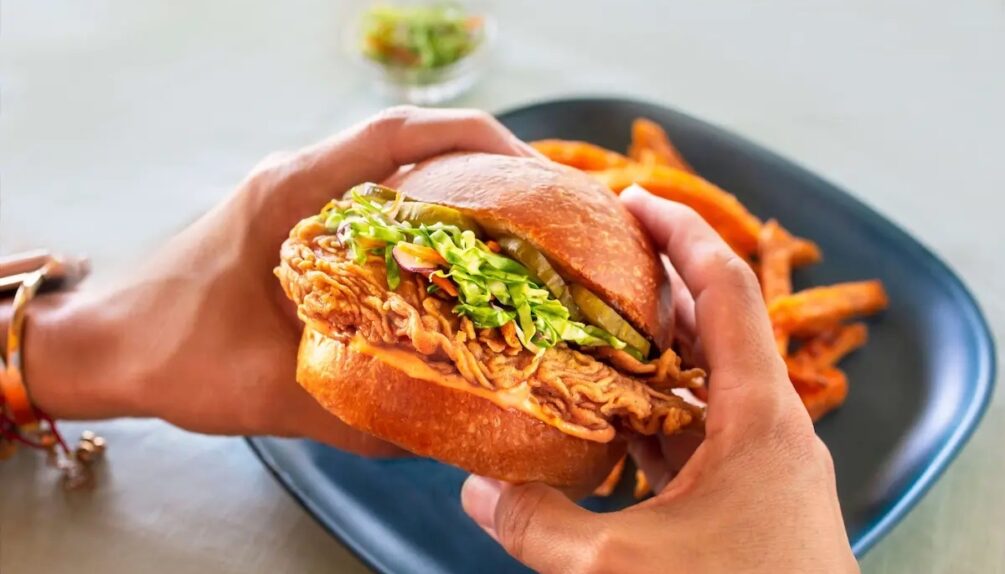 Fried chicken sandwich in bread bun held in hands above blue plate with chips