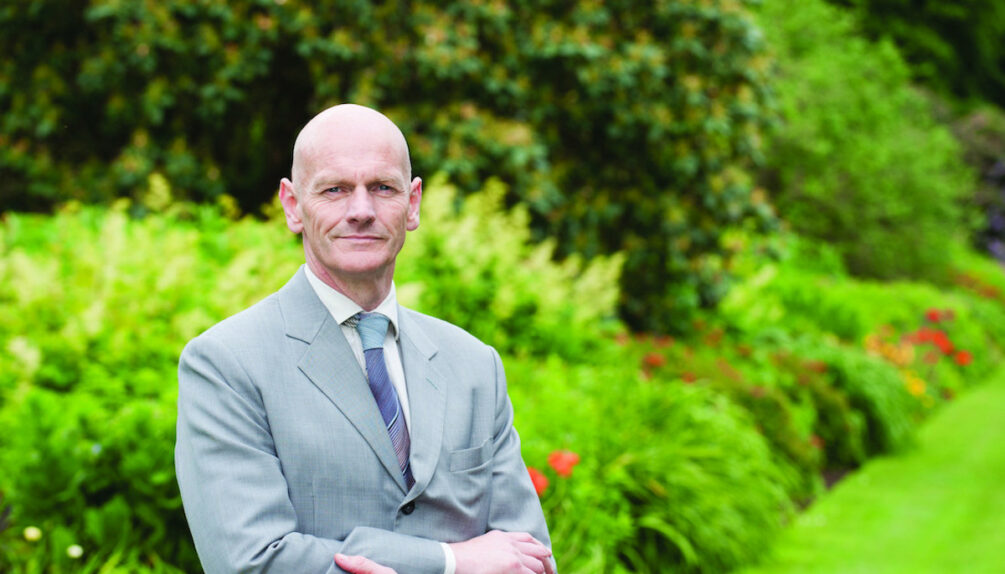 Image of bald man in grey suit in front of greenery background.