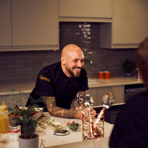 Chef Stu Henshall leaning on kitchen table discussing recip with observer