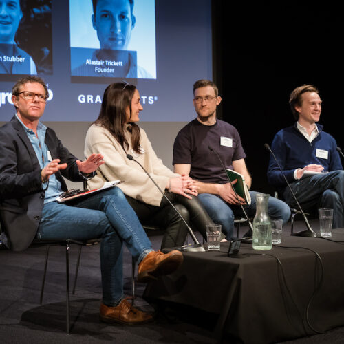Four podcast contributors sat on a stage with a black background, table in front with mics and water glasses